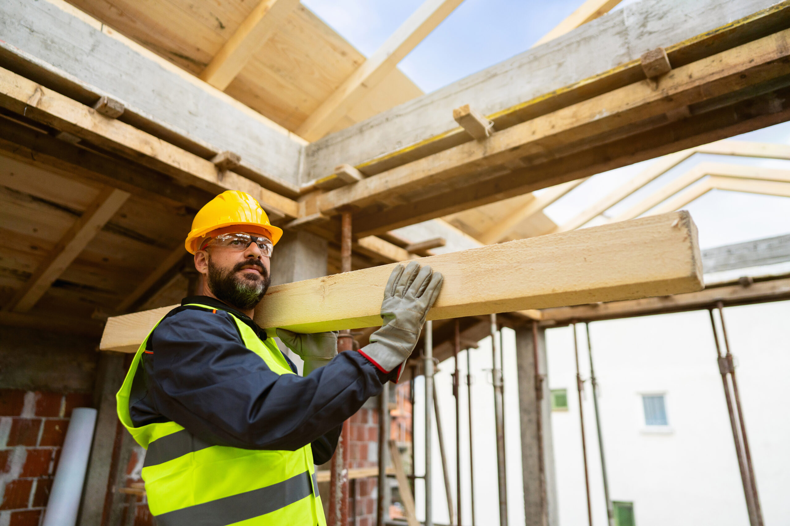 Caucasian male roofer, carrying an timber, while working on a construction site Mid-adult male Caucasian roofer, carrying a big timber, while working on a rooftop, making a roof beam